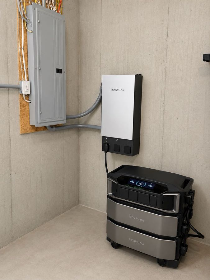 Wall-mounted battery inverter connected by conduit to the service panel, with the battery stack placed alongside
